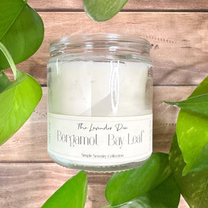 Candle in a clear glass jar with description of bergamot and bay leaf. Candle is sitting on a light wooden surface surrounded by green foliage. 