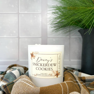 Candle in a white jar with description of Dewey's Snickerdew Cookies scent. Candle is resting on a winter plaid fabric with a snowy day visible in the background.