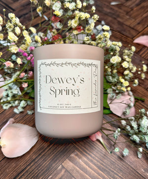Mauve colored candle container with scent label 'Dewey's Spring' resting on a wooden surface surrounded by colored baby's breath and flower petals. 