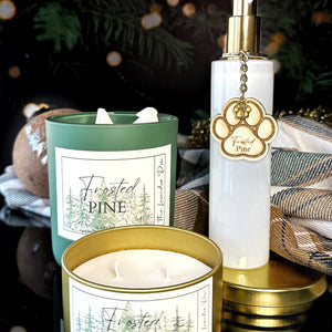 Fragrance collection with scent name of Frosted Pine. One candle is a in dark green jar, one is in a gold metal tin, and the room spray is in a white glass bottle. All items are resting on a glossy black surface with twinkle lights in the background. 