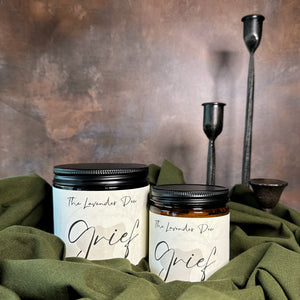 Two candles of different sizes in glass amber jars with black metal lids. The scent name is Grief. The candles are resting on olive green fabric with a metal tinged background.