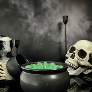 Dyed green candle in a black cauldron shaped container in front of a foggy background and skeleton accents. 