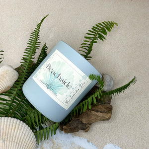 Light blue candle container with scent label 'Beachside' on piece of driftwood with fern and seashell accents. 