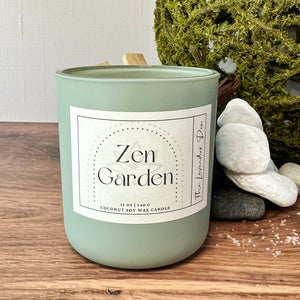 Sage green candle container with scent name 'Zen Garden' on a wooden surface. The backdrop features a large moss ball, some white rocks, and a sprinkle of salt.