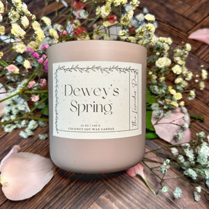 Mauve colored candle jar with scent name Dewey's Spring on a wooden surface surrounded by colored baby's breath flowers and pink petals. 
