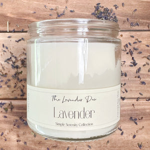 Candle in a clear glass jar with description of lavender. The candle is on a light wooden surface surrounded by lavender buds.