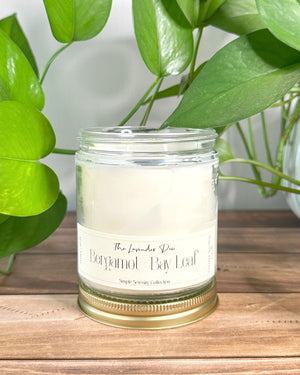 Candle in a glass jar with a label on a wooden surface, surrounded by green leaves.