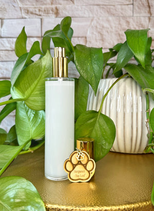 Clear bottle room spray with gold cap and paw-shaped charm on a gold surface with green plants in the background
