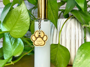 Clear bottle room spray with gold cap and paw print charm on a wooden surface with green plants in the background