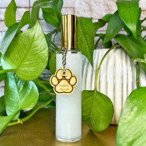 Clear bottle room spray with gold cap and paw print charm on a wooden surface with green plants in the background