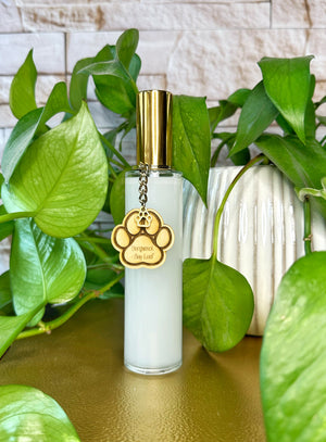 Clear bottle room spray with gold cap and paw print charm on a wooden surface with green plants in the background