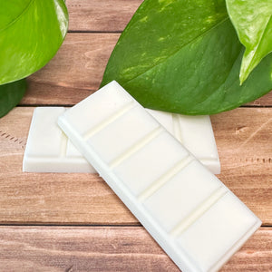 Two white wax melt bars on a wooden surface with green leaves in the background