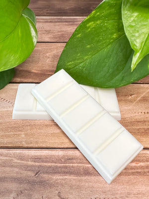Two white wax melt bars on a wooden surface with green leaves in the background