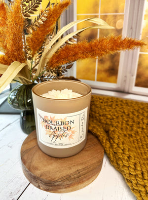 Candle labeled 'Bourbon Braised Apples' on a wooden coaster with decorative elements.