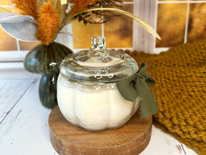 Pumpkin shaped glass jar candle with lid on a wooden stand, surrounded by decorative elements including pampas grass and a textured rug.
