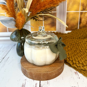 Pumpkin shaped glass jar candle with lid on a wooden stand, surrounded by decorative elements including pampas grass and a textured rug.