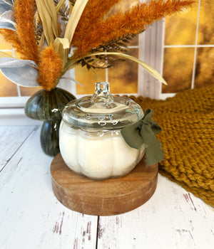 Pumpkin shaped glass jar candle with lid on a wooden stand, surrounded by decorative elements including pampas grass and a textured rug.