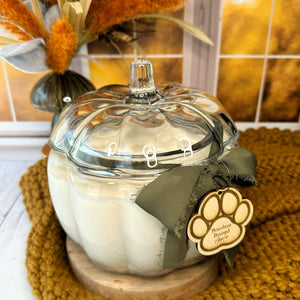 Pumpkin shaped glass jar candle on a wooden coaster with autumn decorations. 
