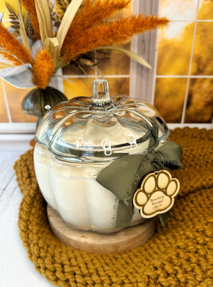 Pumpkin shaped glass jar candle on a wooden coaster with autumn decorations. 