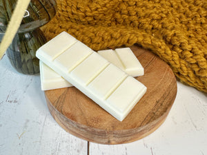 Two white bars of wax melts on a wooden round tray with a knitted yellow blanket in the background.