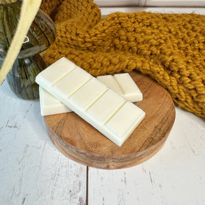 Two white bars of wax melts on a wooden round tray with a knitted yellow blanket in the background.