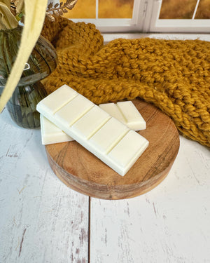 Two white bars of wax melts on a wooden round tray with a knitted yellow blanket in the background.
