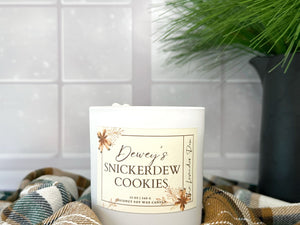 Candle labeled 'Dewey's Snickerdew Cookies' on a checkered fabric with a snowy background