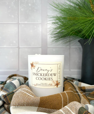 Candle labeled 'Dewey's Snickerdew Cookies' on a checkered fabric with a snowy background