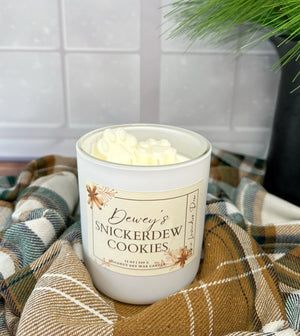 Candle labeled 'Dewey's Snickerdew Cookies' on a plaid fabric surface with  s snowy background.