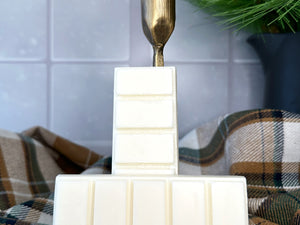 Two creamy white snap bar wax melts leaning against a gold metal candlestick holder. The wax melts sit on a wooden coaster surrounded by a winter fabric and winter decorations. The background is a snowy day visible through a window.