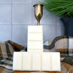 Two creamy white snap bar wax melts leaning against a gold metal candlestick holder. The wax melts sit on a wooden coaster surrounded by a winter fabric and winter decorations. The background is a snowy day visible through a window.