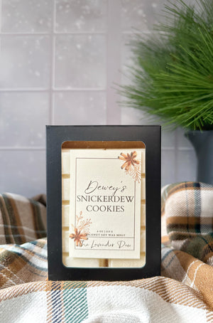Two snap bar wax melts in a black box with a decorative label featuring the scent name: Dewey's Snickerdew Cookies. The box sits on top of a winter plaid fabric with a snowy winter day in the background. 