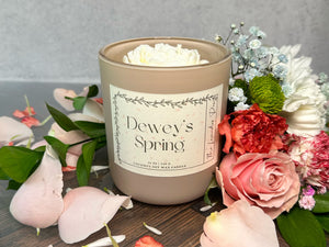 Candle labeled 'Dewey's Spring' surrounded by flowers on a wooden surface