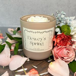 Candle labeled 'Dewey's Spring' surrounded by flowers on a wooden surface