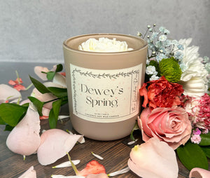 Candle labeled 'Dewey's Spring' surrounded by flowers on a wooden surface