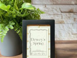Cream colored wax melt snap bars in a black box. The box has a label on it with the scent name of 'Dewey's Spring'. The box and wax melts are sitting on a wooden surface with a plant and white brick in the background.
