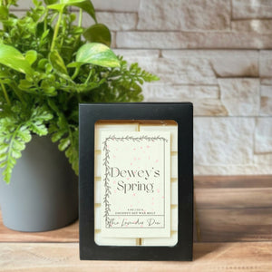 Cream colored wax melt snap bars in a black box. The box has a label on it with the scent name of 'Dewey's Spring'. The box and wax melts are sitting on a wooden surface with a plant and white brick in the background.