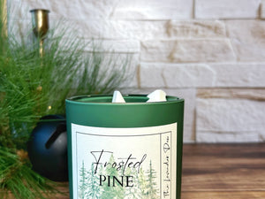 Green candle labeled 'Frosted Pine' on a wooden surface with a stone wall background