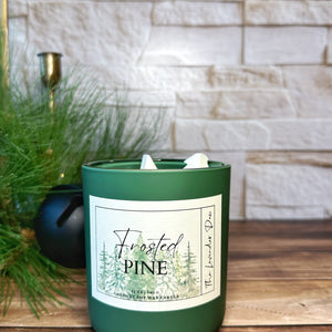 Green candle labeled 'Frosted Pine' on a wooden surface with a stone wall background