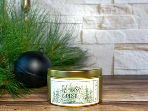 Gold metal tin candle labeled 'Frosted Pine' on a wooden surface with decorative elements.