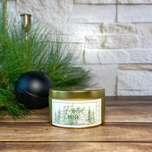 Gold metal tin candle labeled 'Frosted Pine' on a wooden surface with decorative elements.
