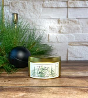 Gold metal tin candle labeled 'Frosted Pine' on a wooden surface with decorative elements.