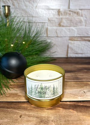 Candle in a gold tin labeled 'Frosted Pine' on a wooden surface with a stone wall background.