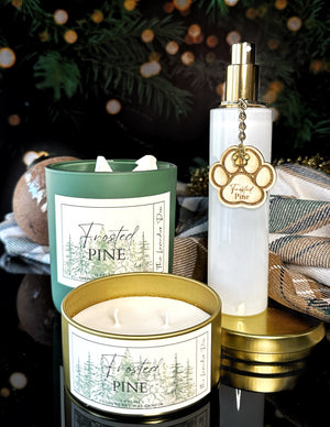 Two candles labeled 'Frosted Pine' and a white and gold room spray bottle on a reflective surface.