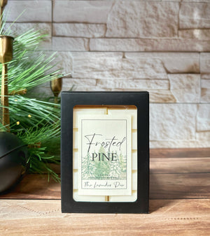 Two wax melt snap bars in a black box with a decorative label featuring the scent name: Frosted Pine. The package sits on a wooden surface with winter decorations and white brick in the background.