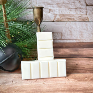 Two creamy white snap bar wax melts leaning against a gold metal candleholder. The wax melts sit on a wooden surface with winter decorations and white brick in the background.