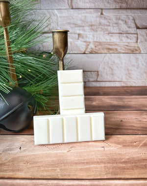 Two creamy white snap bar wax melts leaning against a gold metal candleholder. The wax melts sit on a wooden surface with winter decorations and white brick in the background.
