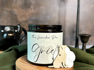Candle labeled 'Grief' by The Lavender Dew on a wooden stand with decorative elements.
