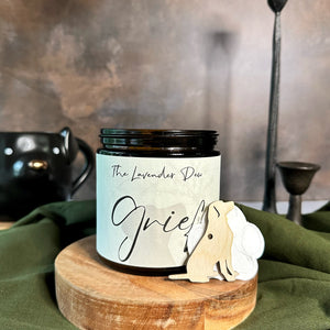 Candle labeled 'Grief' by The Lavender Dew on a wooden stand with decorative elements.