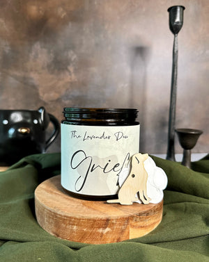 Candle labeled 'Grief' by The Lavender Dew on a wooden stand with decorative elements.